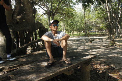 Portrait of young man sitting on wood against trees