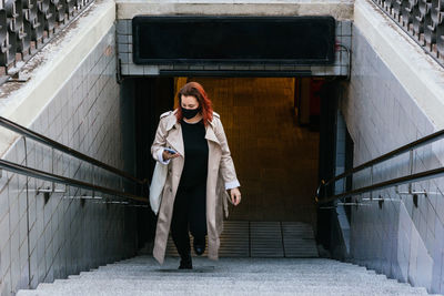 Female in protective mask walking upstairs in city and browsing mobile phone during coronavirus epidemic