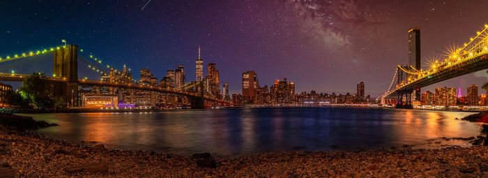 Illuminated bridge over river by buildings against sky at night