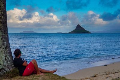 Side view of woman looking at sea while sitting on shore against tree trunk