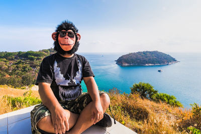 Young man sitting by sea against sky