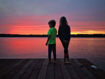 Rear view of friends standing by lake against sky during sunset