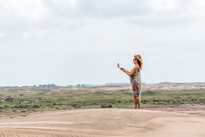 Blonde woman in a colourful summer dress standing on a sand dune taking pictures with her mobile