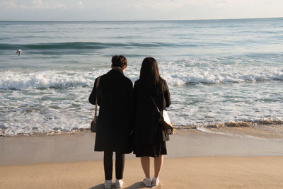 Rear view of couple standing on shore at beach against sky