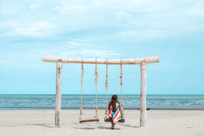 Full length of woman sitting on swing at beach against sky