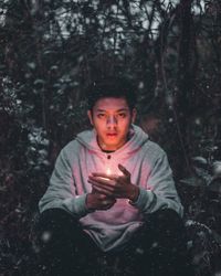 Portrait of young man holding burning candle while sitting amidst plants during winter