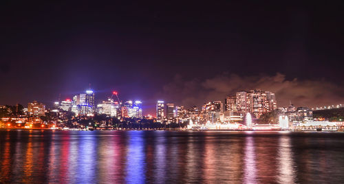Illuminated buildings by river against sky at night