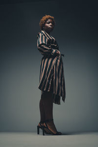 From below full body of self assured black female model in striped dress and high heels keeping hands in pockets while standing in dark studio