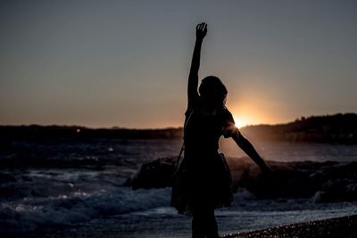 Silhouette woman with hand raised standing at beach against sky during sunset