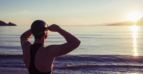 Back view of unrecognizable swimmer in swimwear and goggles standing on seashore and enjoying sunset during training