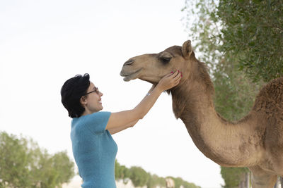 Side view of woman touching camel standing against clear sky