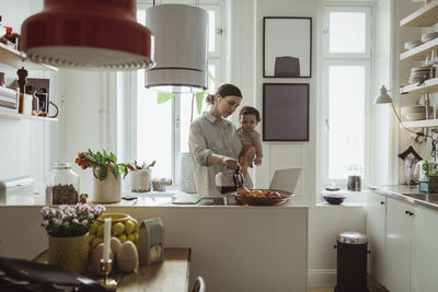 Female professional pouring coffee while carrying toddler son in kitchen