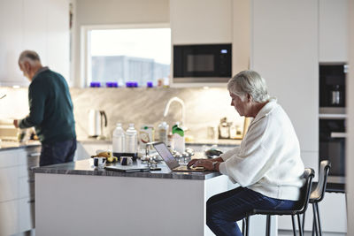 Senior woman using laptop while male preparing food in kitchen