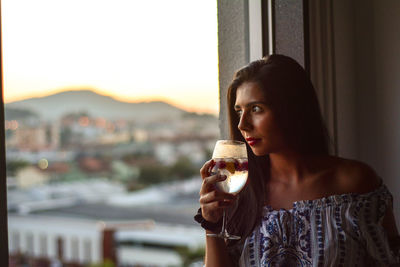 Thoughtful young woman drinking wine while looking through window