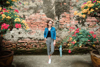 Woman in sunglasses standing amidst flowering plants against brick wall