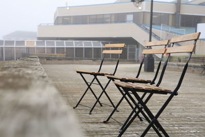 Empty chairs and table on beach against buildings in the fog