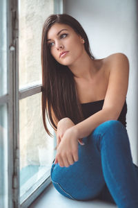 Thoughtful young woman looking through window while sitting at home