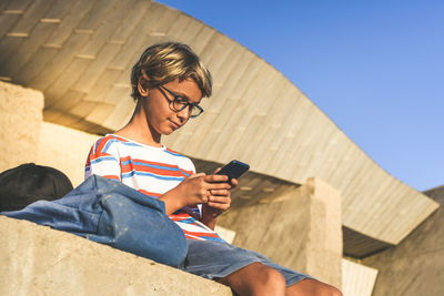 Low angle view of boy using phone while sitting outside building