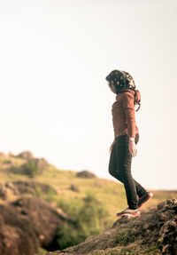 Side view of young woman standing on rock against clear sky