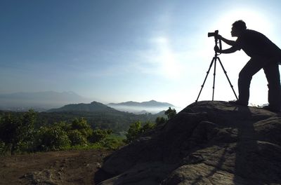 Silhouette man photographing with camera and tripod on mountain against sky