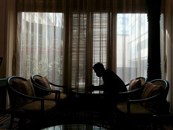 Side view of man sitting on chair by covered window at home
