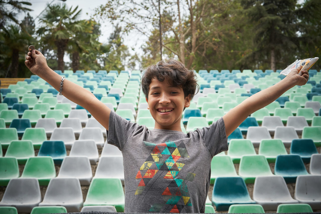 smiling, childhood, portrait, child, one person, happiness, front view, looking at camera, men, arm, emotion, limb, cheerful, standing, human limb, day, arms raised, waist up, nature, green, enjoyment, tree, plant, fun, casual clothing, arms outstretched, person, outdoors, leisure activity, teeth, smile, lifestyles