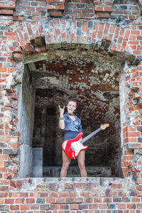 Portrait of female model standing with guitar at window amidst damaged brick wall