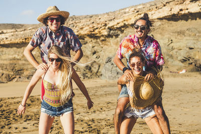 Girlfriends piggybacking boyfriends at beach against sky