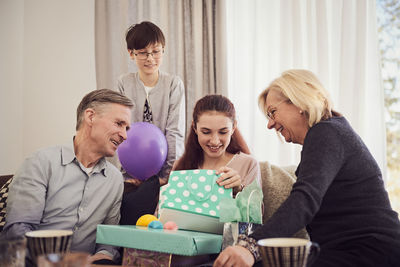 Family looking at smiling girl opening gifts on sofa at home