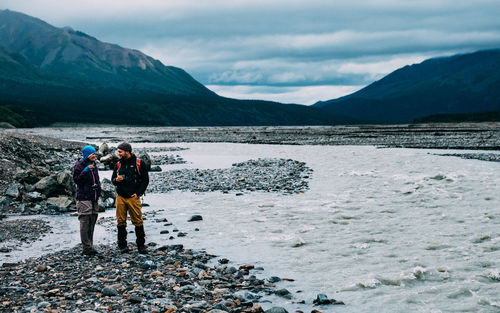 Friends standing at lakeshore against mountains
