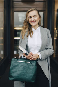 Portrait of businesswoman with book and bag standing by store in city