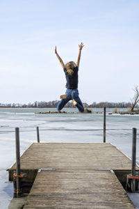 Rear view of woman jumping on pier over lake against sky