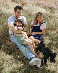 Side view of family with father, two daughters and small black dog sitting on field and smiling 