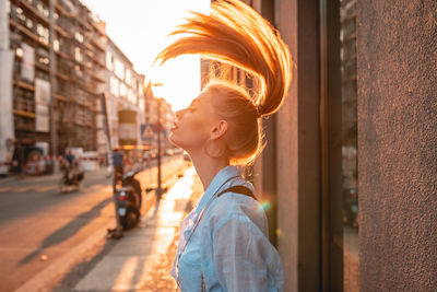 Side view of young woman tossing hair while standing on sidewalk during sunset