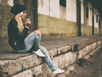 Full length side view of young woman smoking cigarette while sitting against building