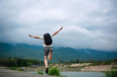 Back view point of young man backpacker jumping over the ground and raising arm with victory sign