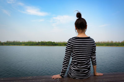 Rear view of woman sitting by lake against sky