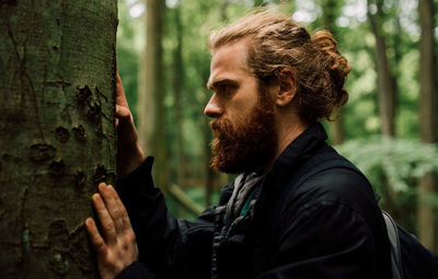 Young man looking away by tree trunk in forest