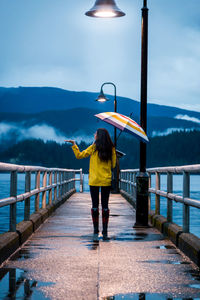 Rear view of woman with umbrella walking on pier over lake against mountains