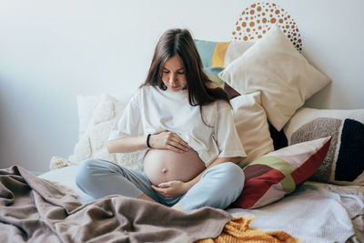 A pregnant woman sitting on the bed in homewear gently holding her pregnant tummy.