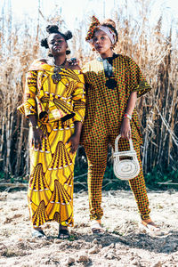 Self assured black female models in stylish clothes with authentic african prints looking at camera while standing together against dry grass in countryside