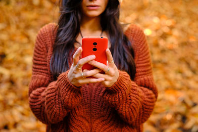 Cropped unrecognizable young female in warm red knitted pullover browsing mobile phone while standing in autumn park with fallen colorful leaves