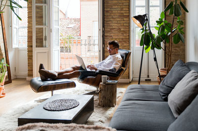 Handsome male entrepreneur sitting in armchair at home and working on project on laptop while enjoying coffee