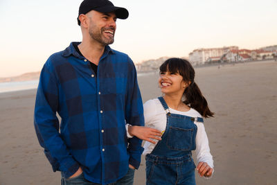 Father and daughter walking arm in arm at beach against sky