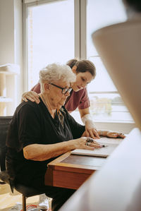 Female caregiver with hand on shoulder of senior woman sitting at desk in apartment