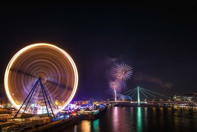 Illuminated ferris wheel by river against sky at night