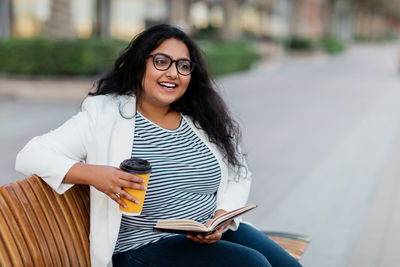 An indian girl is sitting on a bench, reading a book and drinking coffee.