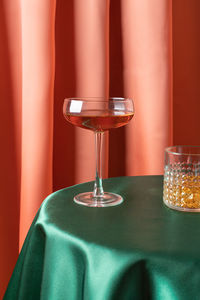 Elegant crystal glasses of rose wine and whiskey on table covered with smooth silk tablecloth against drapery fabric