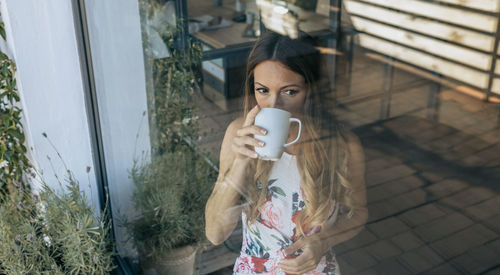 Woman looking away while drinking coffee seen through glass window