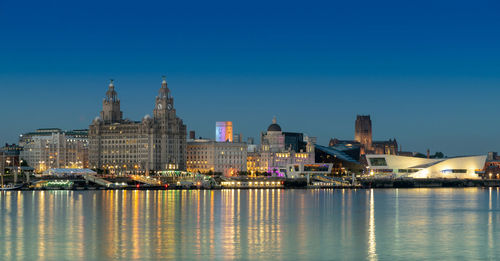 Liverpool skyline, illuminated buildings by river against clear blue sky